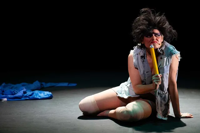 Dynasty Handbag sits on a dark stage in a torn denim vest, thigh-high stockings, and voluminous black wig, holding an oversized yellow pencil to her mouth like a microphone. A crumpled blue garment lies on the otherwise bare stage behind her.