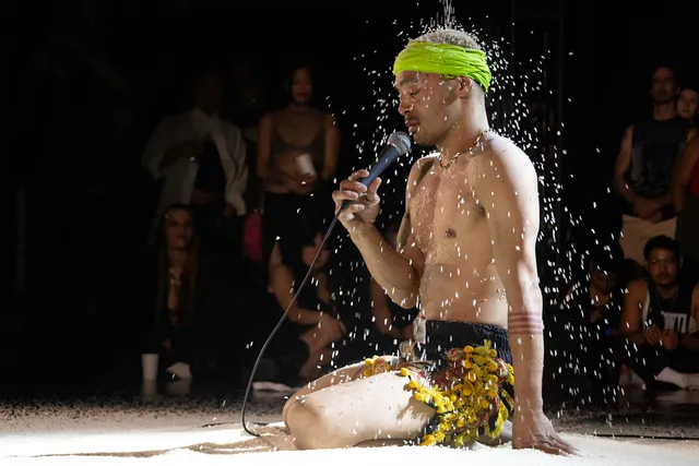 A shirtless performer kneels on a sandy stage, speaking into a microphone while a shower of sand or small particles falls around them. The audience, visible in the background, watches intently.