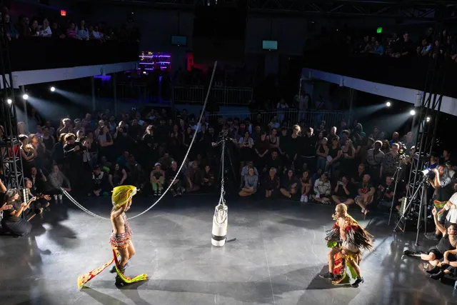 Two performers in colorful costumes face each other in a ring surrounded by a large audience under dramatic lighting in an indoor arena.