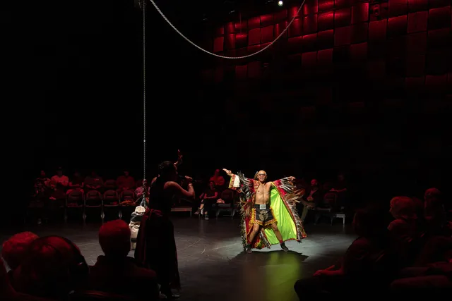 a filipino performer in a indigenous cape with chicken feathers spreads his arms wide near a hanging punching bag surrounded by audience in 360 degrees.