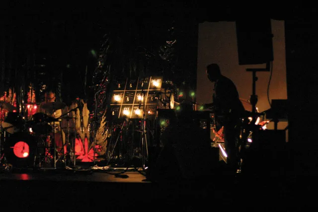 A man silhouetted on a dark stage cluttered with various colored lights, a keyboard, and a drum kit. 