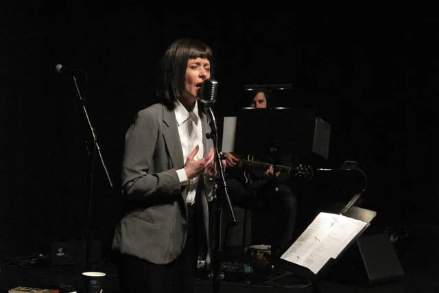 A woman wearing a grey suit with a white button down shirt sings with a heartfelt expression into a microphone on a dark stage. 