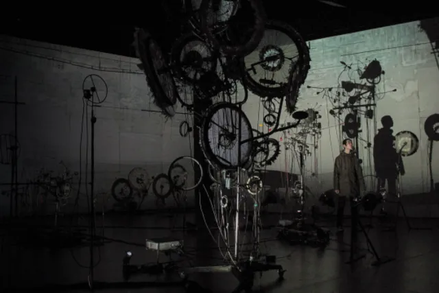 A man casting a long shadow standing near a sculpture of gears and wheels in a grey room. 