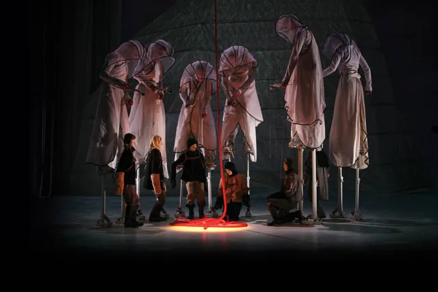 Puppeteers wearing white hooded cloaks, standing on stilts while controlling marionettes of people gathered around a red rope. 