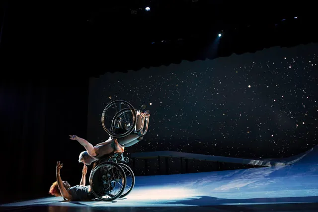 A woman in a wheelchair flying in the air with arms spread wide, wheels spinning, supported by another woman in a wheelchair who is lifting from the ground below.  