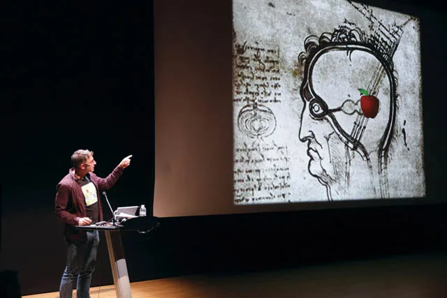 A man standing on a dimly lit stage gesturing at a projection of a drawing by Leonard Da Vinci with an emoji apple. 