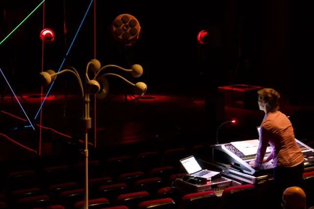 A woman standing behind an audio mixer looking at projections of red, blue, purple, and yellow 3D lines and shapes on a dark stage. 