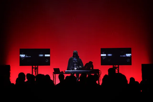 A DJ wearing a cloak playing to a silhouetted audience infant of a wall of red light. 