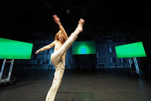 A dancer wearing a cream colored outfit kicking, with leg and arm in the air and blurred in motion. Three green screens are postponed around them in a black box studio. 