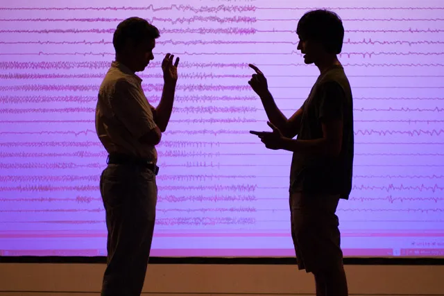 Two men silhouetted in front of a purple projection of sound waveforms. 