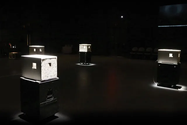 White cubes with small windows placed through a dark space on black pedestals. 