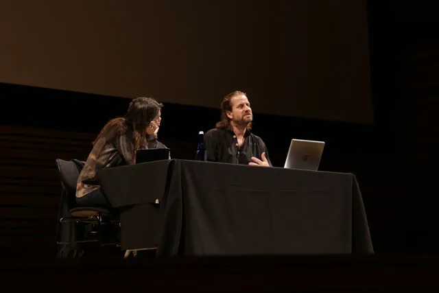 A man and woman sitting in front of a black table on a dark stage in discussion. 