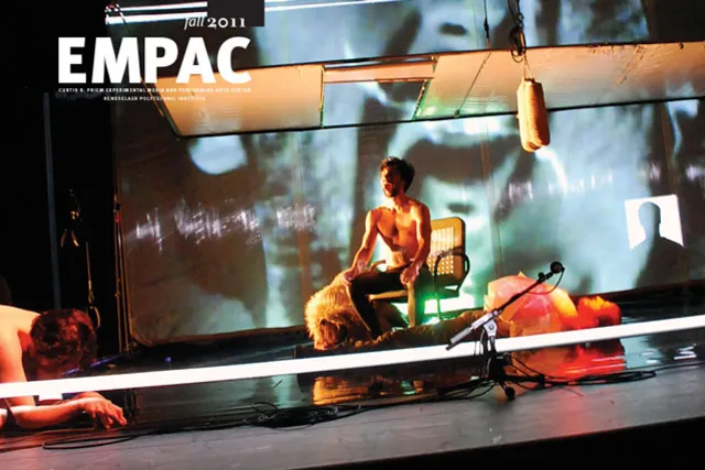 A shirtless white man sitting in a chair screaming as other performers lay on the stage around him lit in orange light, EMPAC Fall 2011. 