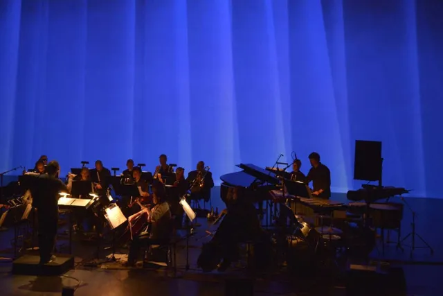 An orchestra performing on a black stage in front of a wall of blue light. 