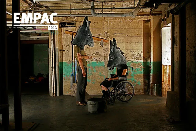 Two people, one standing and one in a wheelchair wearing donkey heads in front of a wall of peeling paint, EMPAC 2013 