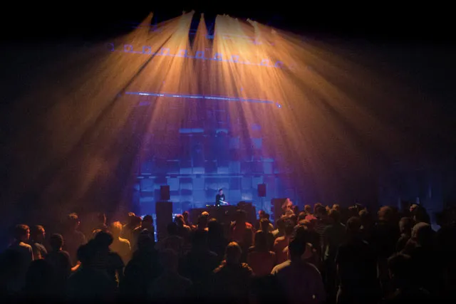 A man DJing to a silhouetted crowd in front of a wall of blue light and yellow spotlights. 