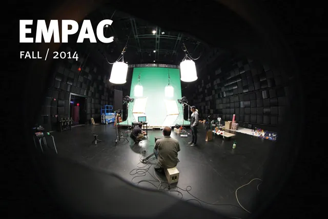 A green screen and studio lighting rigs set up in a black box studio as there crew members look on, EMPAC Fall/2014 in bold white font. 