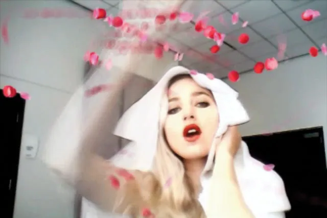 A white woman with bleach blonde hair and red lipstick throwing red rose petals at the camera with a white cloth over her head. 