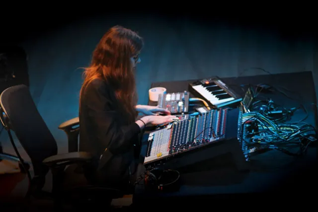 A white woman sitting behind a sound board and keyboard in a blue tinged room. 