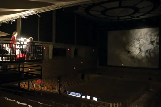 Two people standing on scaffolding in the back of the concert hall working a projector, projecting and image of a dense gray smoke plume. 