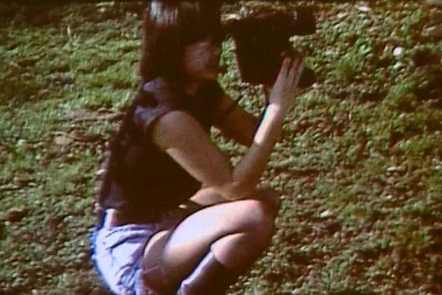 An asian woman from the 90's squatting in the grass with a VHS camera 