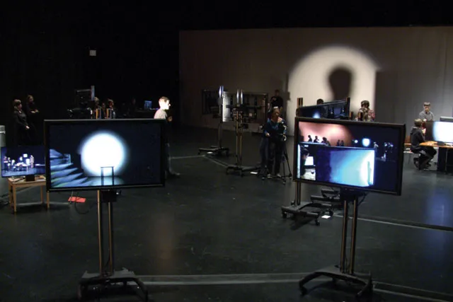 Two screens on legs in for the foreground showing images of the rest of the room. Various crew members work behind the scenes in front of a wall lit by a soft circle of white light. 