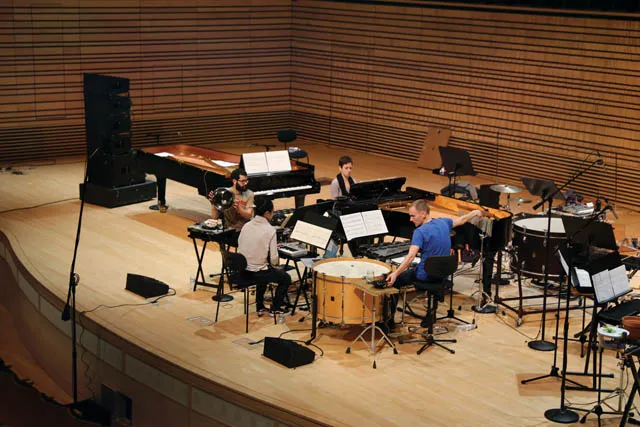 A small orchestra in rehearsal on the concert hall stage surrounded by various percussion and pianos. 