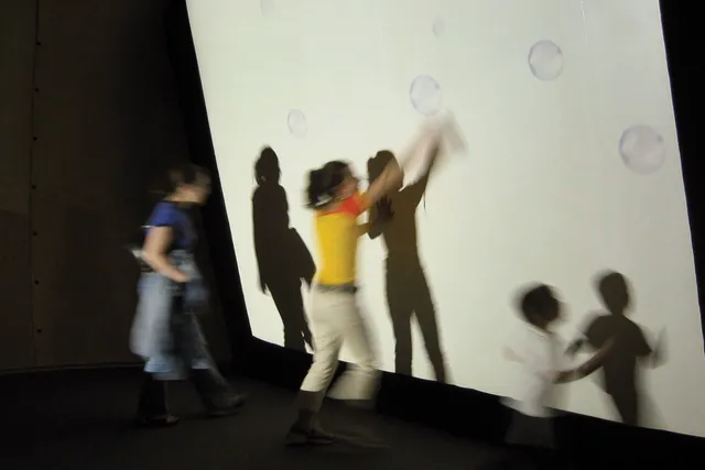 Three children blurred in motion trying to catch projected bubbles on a white wall. 