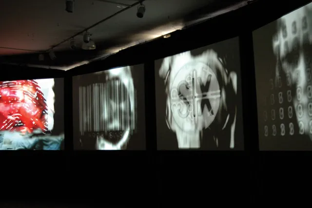 Four large screens on a dark background projecting three abstract black and white images and one of an up close bloody fingerprint. 