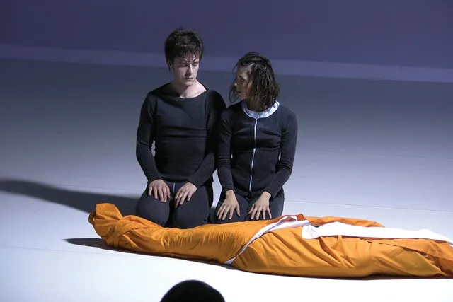 Two people dressed in black unitards kneeling in front of a body wrapped in orange cloth. 
