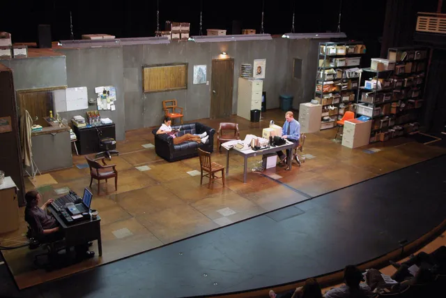 A set of a cluttered office space on a stage. A man wearing business casual sits at a table in the middle of the scene at an old computer. A woman is lounging on a black couch behind him. 