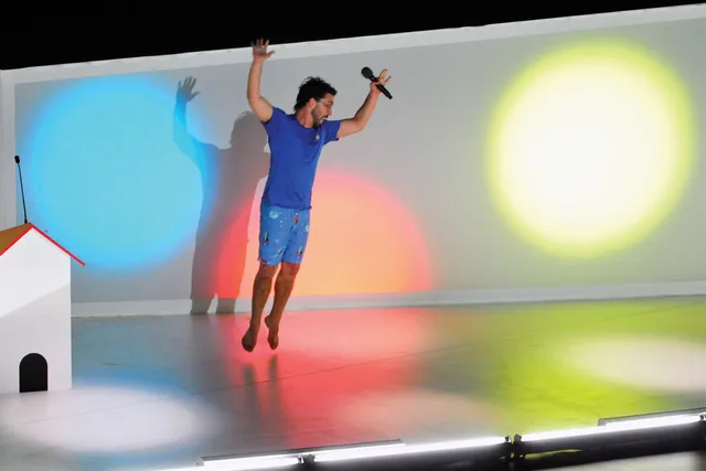 A man wearing a blue t-shirt and shorts jumping with arms up in a white room lit with primary colored lights. 