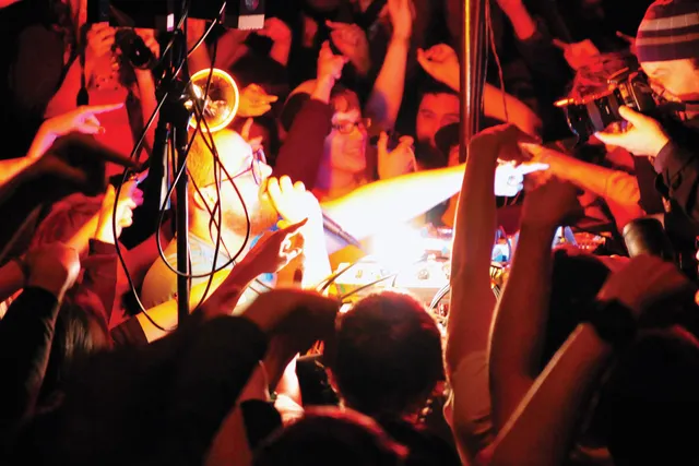 Dan Deacon performing in the middle of a tightly packed crowd, all pointing in the same direction. A beam of yellow light over exposes the middle of the scene while the rest is lit in red light. 