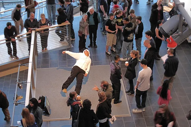 People gathered in the mezzanine watching a performer wearing a white hoodie and black pants leaning back dramatically, almost falling over. 