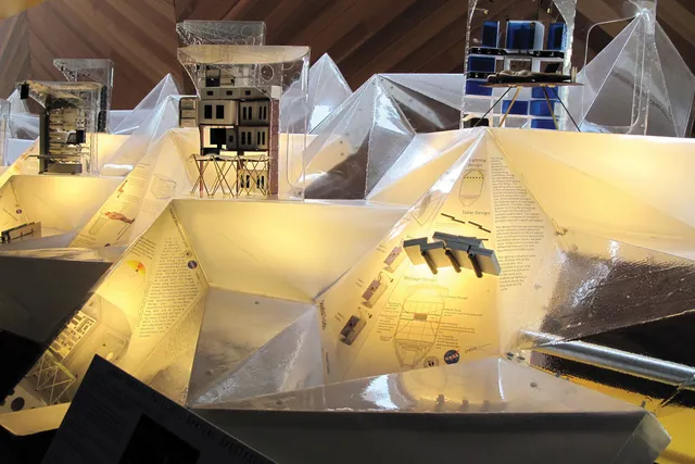 Transparent and frosted pyramids lit internally by yellow light. Scaffolding holding various cubes are placed throughout the pyramids. 