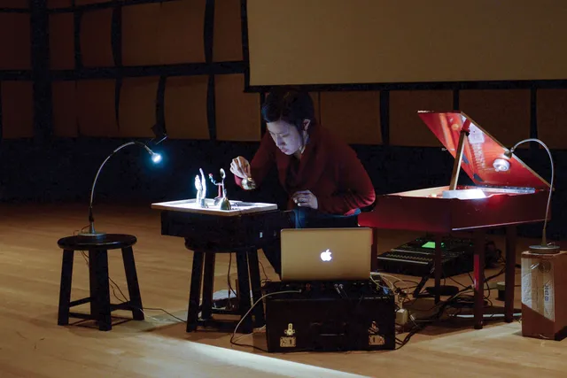 A woman hunched at a small desk setting up figurines under a light. She is surrounded by a small red piano and sound recording equipment. 