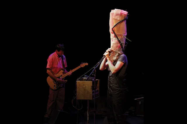 A person singing into a microphone while wearing an abstract tall headdress of wicker canning, zip ties, and a bike tire. 