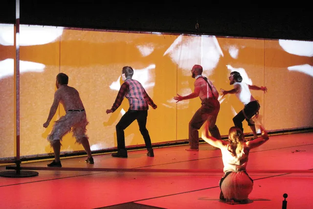 Four male performers yellowing at a yellow wall with outstretched arms. A woman sits on her knees on a red floor in the foreground with back to the viewer with her arms up, directing the performance. 