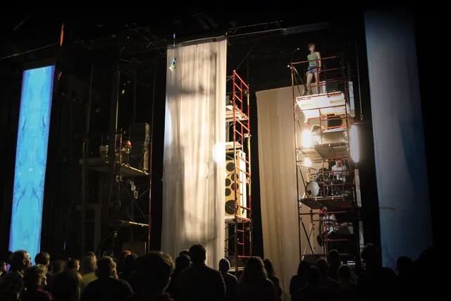 Musicians on scaffolding with each member of the band on different levels playing a concert to a crowd on a staged draped in sheer white fabric. 