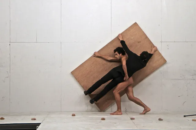 A nude man carrying another man wearing a black unitard while both hold a large piece of plywood on a white stage. 