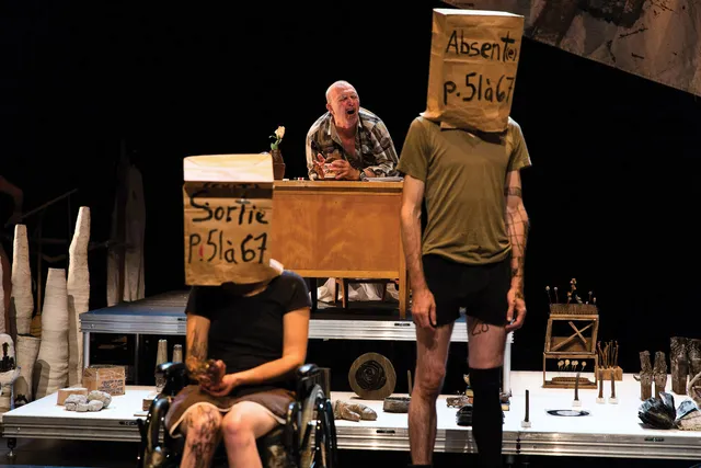 Three people on a cluttered stage. A white woman seated in a wheelchair and a white man standing in his underwear both wearing paper bags over their head. The third man sits behind them at a wooden desk yelling at the couple. 