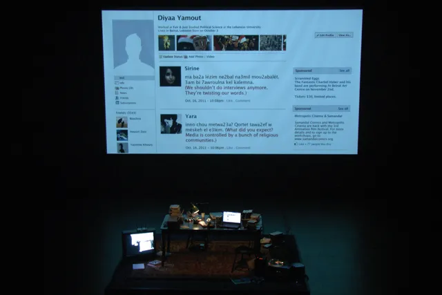 A cluttered desk on a black stage in front of a large screen projecting an image of an old Facebook profile. 