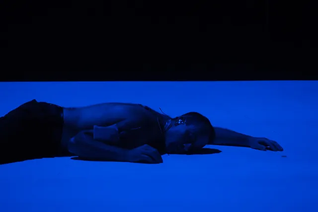 A male performer laying on stage washed in blue light. 