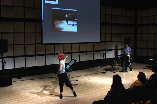 Frédéric Bevilacqua giving a lecture in front of a wall of gray acoustic tiles as a female dancer wearing a red winter hat moves across the stage.