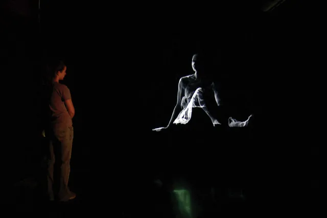 A woman studying the projected image of a ghostly woman seated with legs out, supported by her arms behind her. 