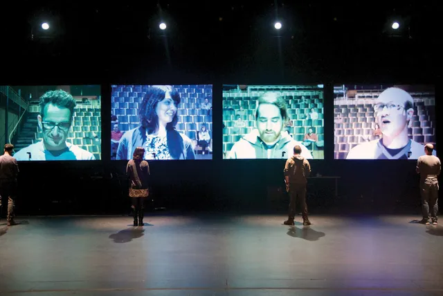Four people standing on stage, back to the viewer looking at a projected image of themselves on a four screens in front of them. 