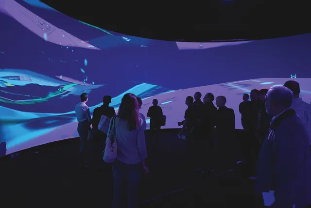 A small audience silhouetted against a panoramic screen showing abstract purple and teal waves. 