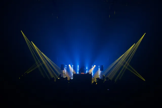 A blue lit stage with yellow spot lights with the silhouette of a DJ and their set up playing in front of a crowd. 