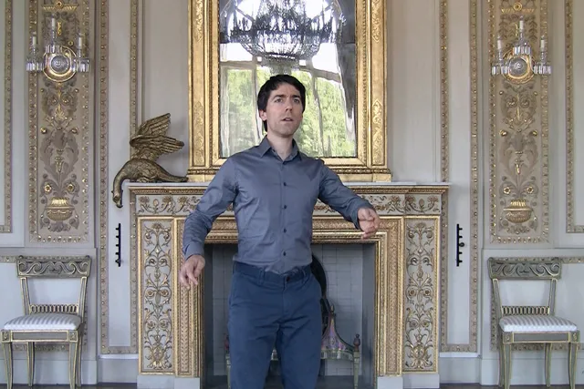 A white man with dark hair wearing a blue button up and pants standing with arms gently gesturing outward in a baroque styled room. 