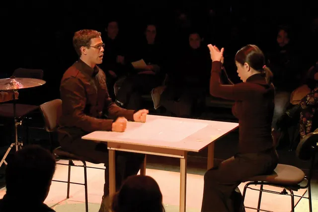 Two people seated at a small square table on a stage. The woman has her hand in a clapping motion above her head. The man looks t hr with mouth open and hands on table in fists. 